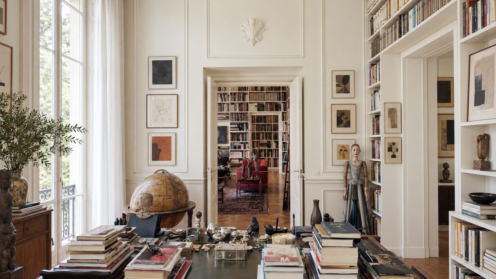 A white-walled study filled with books, drawings and collected objects, opening onto a library